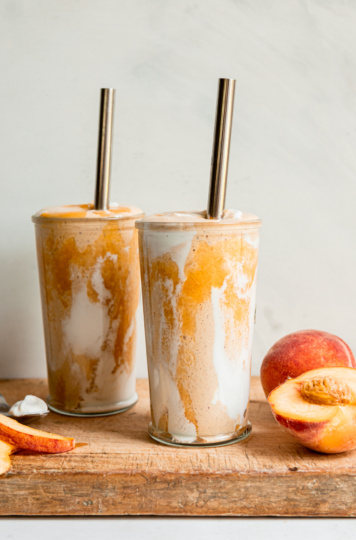 A head-on shot of a vegan peaches and cream protein smoothie in two glasses with metal straws. Peach purée and vegan yogurt create a tie dye effect on the glass.