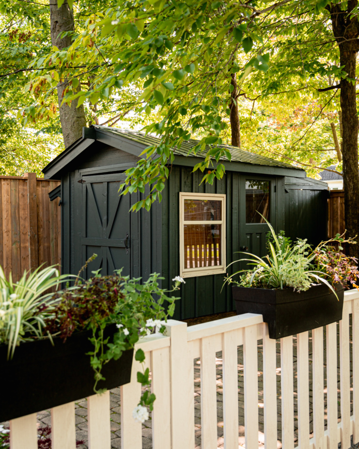 A head-on shot shows a dark garden shed with wood siding, surrounded by trees. There is a fence with planters in front of the shed.