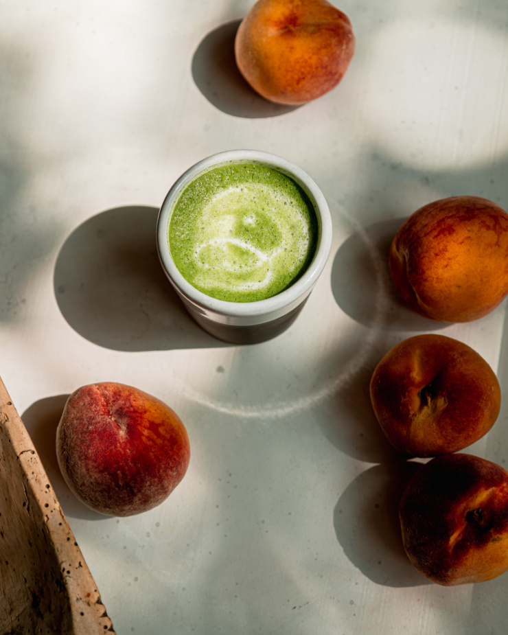 An overhead shot shows a small, frothy matcha latte and a few peaches nearby.