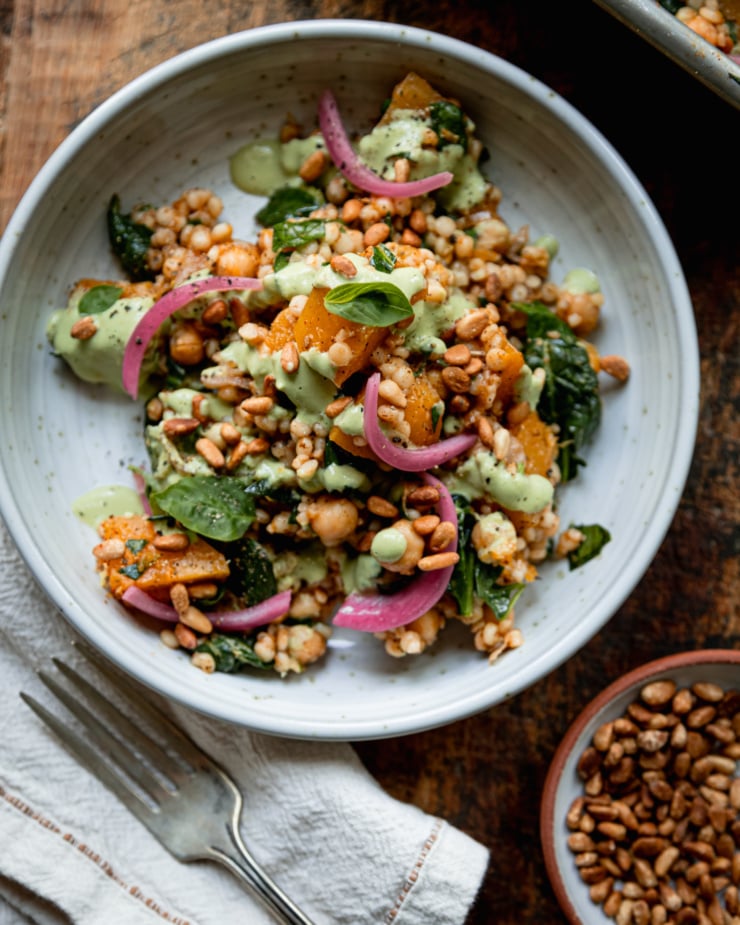 An overhead shot shows an individual serving of baked pearl couscous and butternut squash in a bowl. The bake features chickpeas and spinach, and is topped with pickled red onions and drizzles of a creamy green basil tahini sauce.
