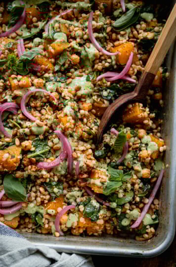 An overhead shot shows a baking dish filled with baked pearl couscous and butternut squash. The bake features chickpeas and spinach, and is topped with pickled red onions and drizzles of a creamy green basil tahini sauce. A wooden serving spoon is sticking out of the dish.