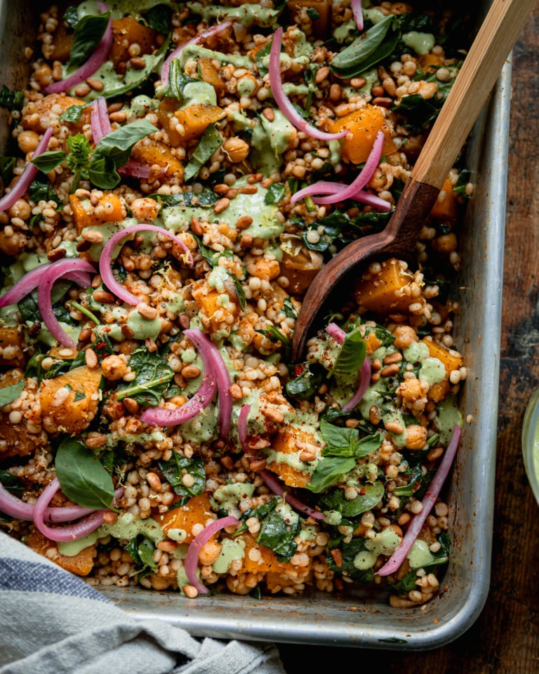 An overhead shot shows a baking dish filled with baked pearl couscous and butternut squash. The bake features chickpeas and spinach, and is topped with pickled red onions and drizzles of a creamy green basil tahini sauce. A wooden serving spoon is sticking out of the dish.