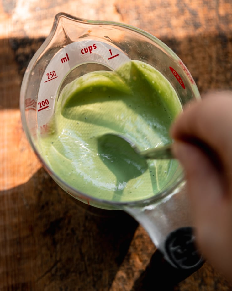 An overhead shot shows a hand using a spoon to stir up some creamy green basil tahini sauce in a measuring cup.