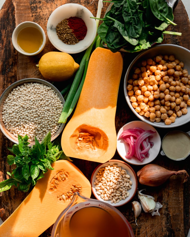 An overhead shot shows ingredients used in a vegan dinner recipe, featuring butternut squash, pearl couscous, pine nuts, chickpeas, spinach, pickled red onions, vegetable stock, shallot, garlic, lemon, basil, olive oil, spices, and green onions.