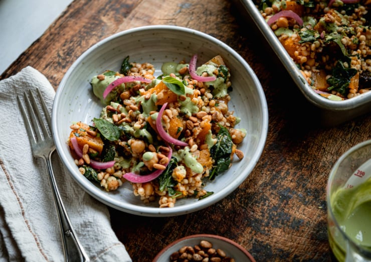 A 3/4 angle shot shows an individual serving of baked pearl couscous and butternut squash in a bowl. The bake features chickpeas and spinach, and is topped with pickled red onions and drizzles of a creamy green basil tahini sauce.