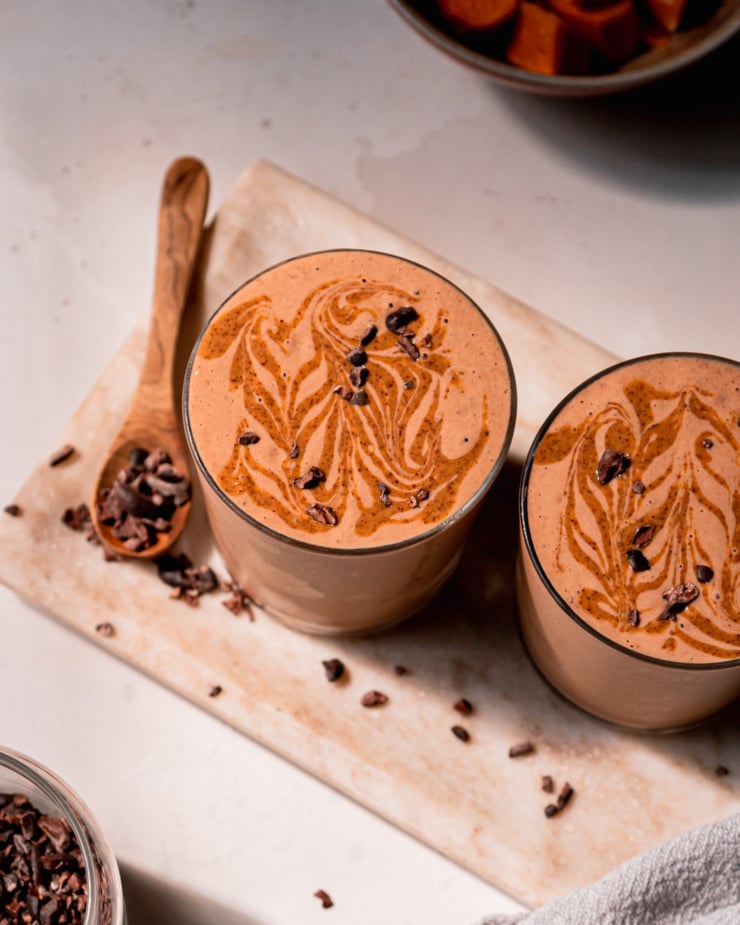 An overhead shot shows two glasses of chocolate sweet potato smoothie with almond butter swirls and cacao nibs on top. The glasses are perched on a beige marble platter.