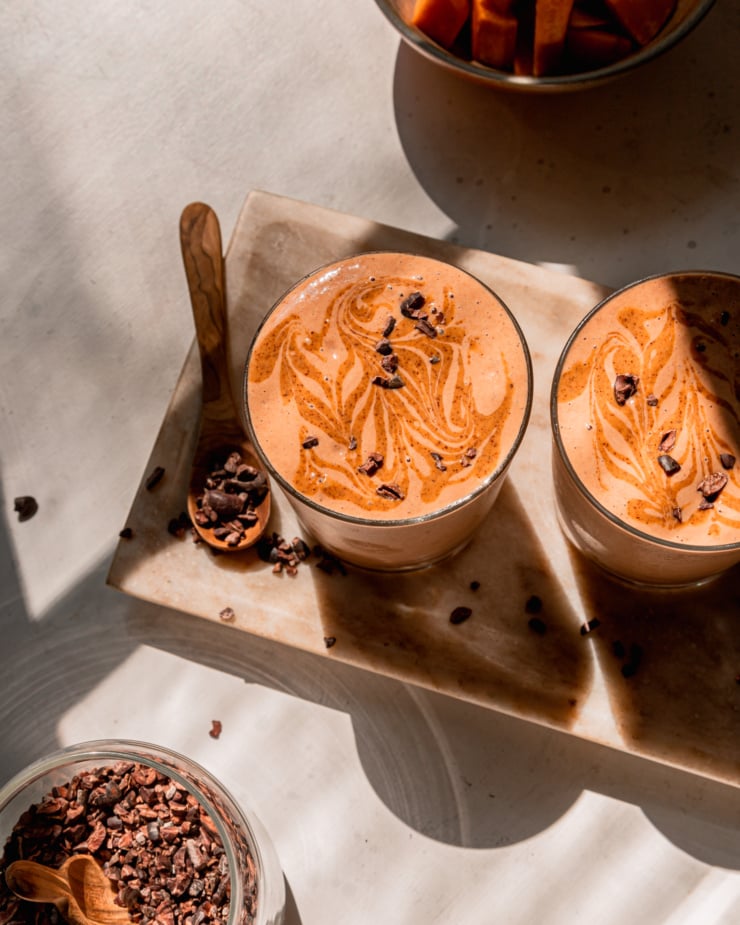 An overhead shot shows two glasses of chocolate sweet potato smoothie with almond butter swirls and cacao nibs on top; all in direct sunlight. The glasses are perched on a beige marble platter.