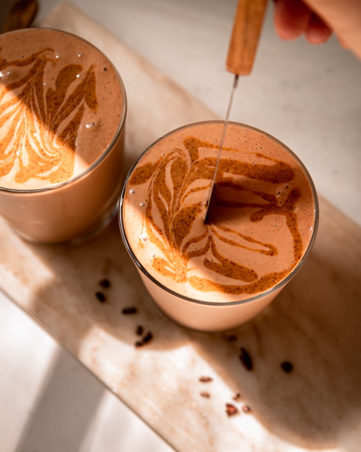 An overhead shot shows a hand using a paring knife to create almond butter swirls in the top of a smoothie.