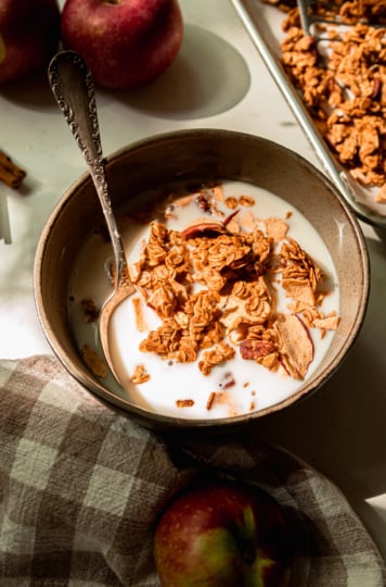 A 3/4 angle shot shows a serving size bowl of apple cinnamon granola with almond milk. A spoon is sticking out of the bowl.