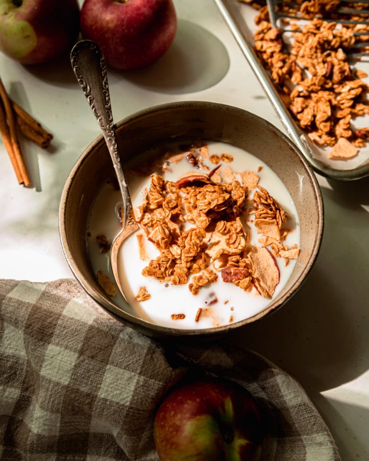 A 3/4 angle shot shows a serving size bowl of apple cinnamon granola with almond milk. A spoon is sticking out of the bowl.