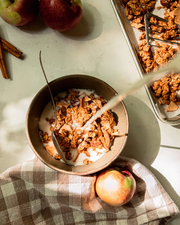 An overhead shot shows almond milk being poured into a bowl of apple cinnamon granola.