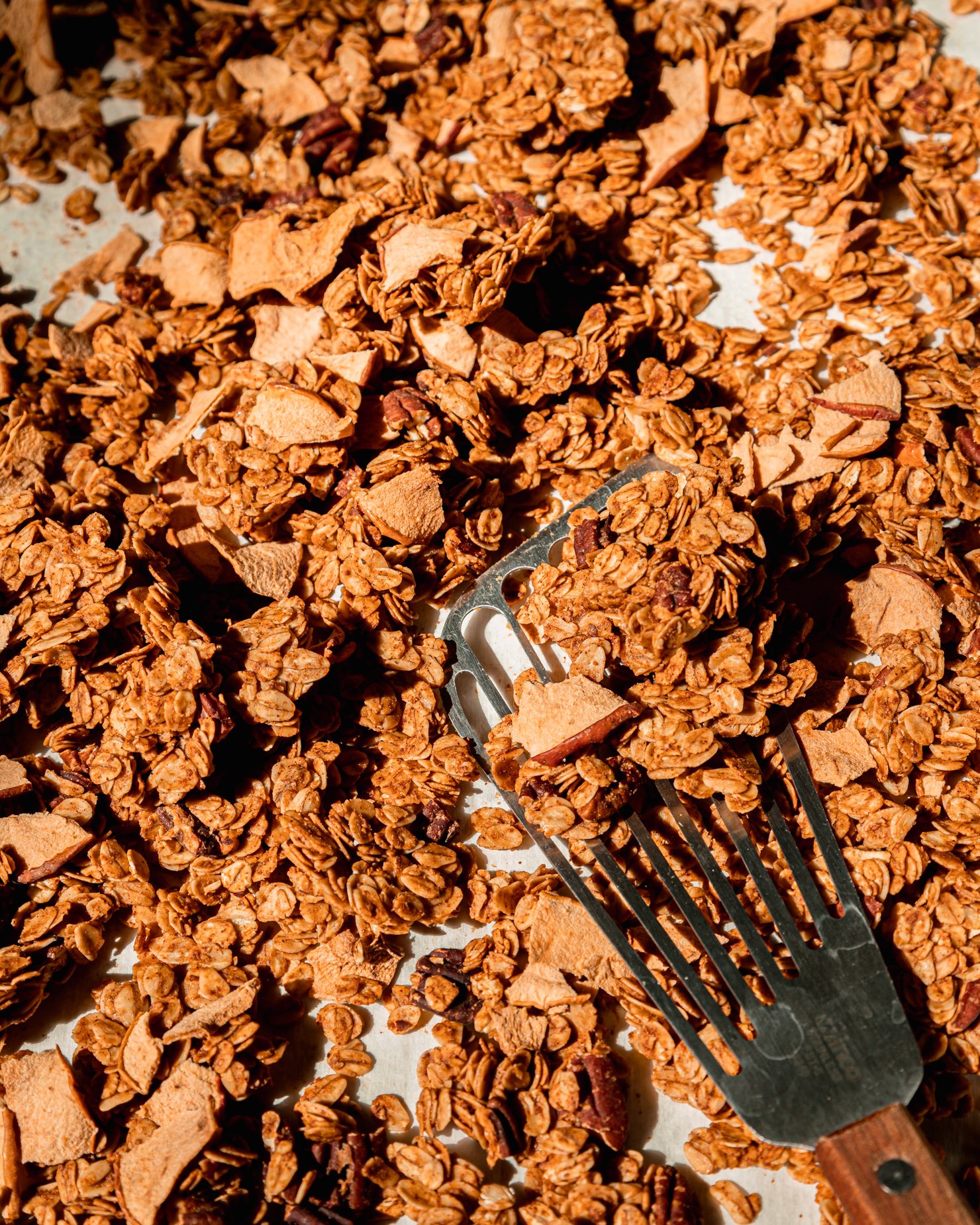 An overhead shot shows broken up clumps of apple cinnamon granola on a sheet pan with a spatula nearby.