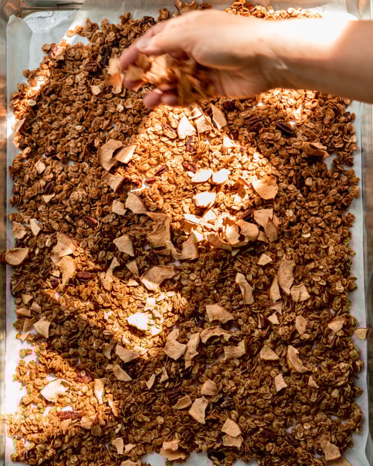 An overhead shot shows a hand scattering pieces of apple chips over a sheet pan filled with apple cinnamon granola.