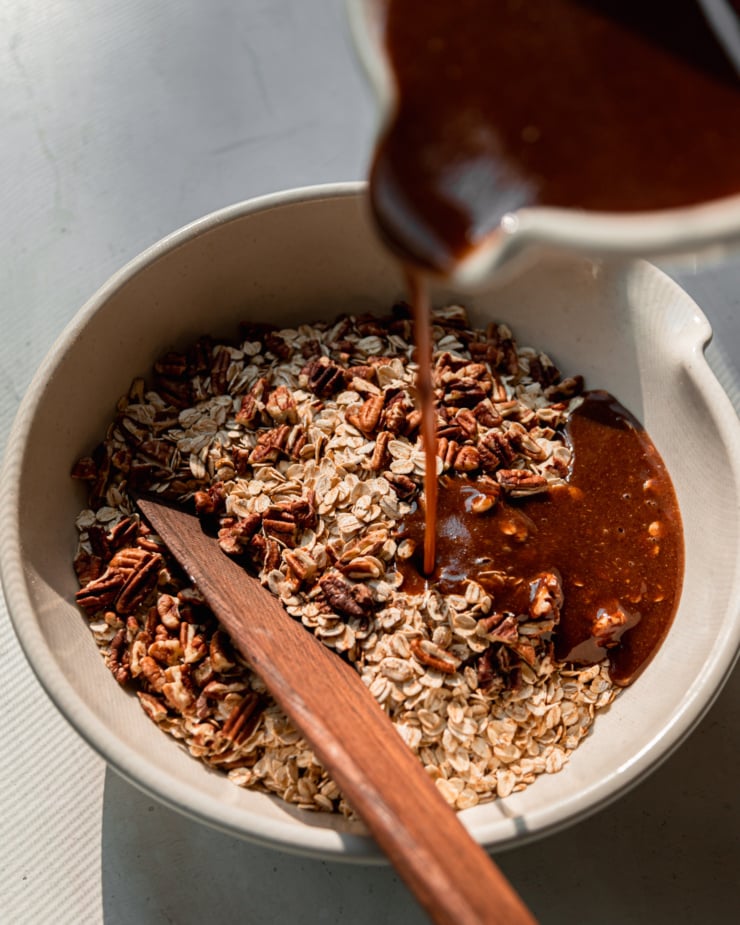 A 3/4 angle shot shows a cinnamon-y liquid mixture being added to a bowl of oats and chopped pecans.