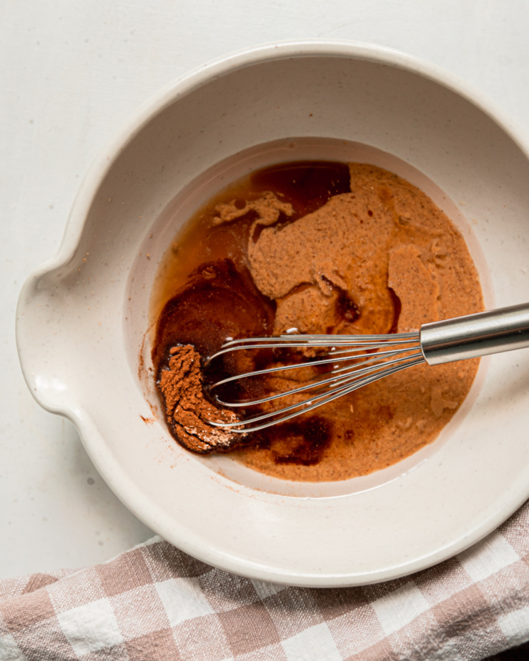 An overhead shot of a mixing bowl with melted coconut oil, almond butter, maple syrup, vanilla, and spices. A whisk is sticking out of the bowl.