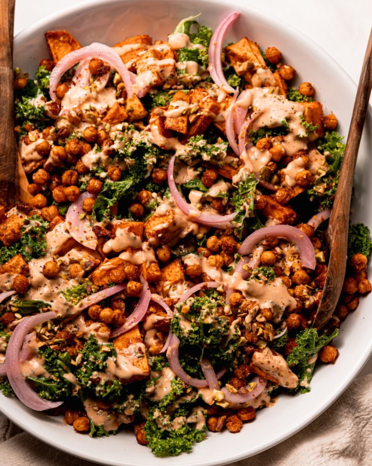 An overhead shot shows a kale sweet potato salad with crispy chickpeas, a creamy and spicy dressing drizzled over top, slices of pickled red onion, and finely chopped pistachios. Wooden salad tons are sticking out of the wide serving bowl.