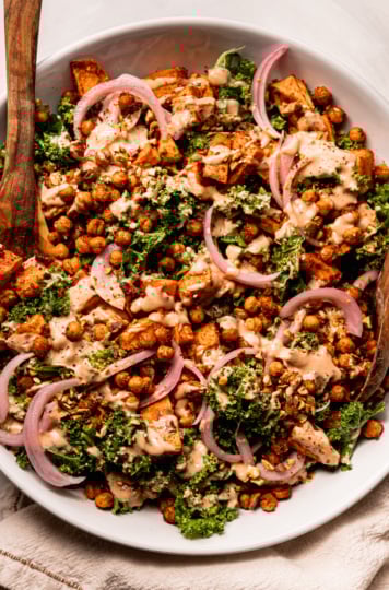 An overhead shot shows a kale sweet potato salad with crispy chickpeas, a creamy and spicy dressing drizzled over top, slices of pickled red onion, and finely chopped pistachios. Wooden salad tons are sticking out of the wide serving bowl.