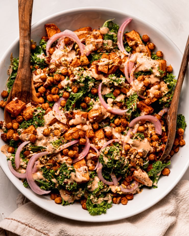 An overhead shot shows a kale sweet potato salad with crispy chickpeas, a creamy and spicy dressing drizzled over top, slices of pickled red onion, and finely chopped pistachios. Wooden salad tons are sticking out of the wide serving bowl.