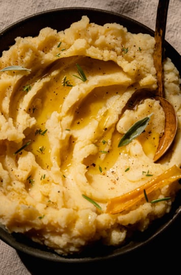 An up close, overhead shot of olive oil mashed potatoes in a bowl with swoops of olive oil on top and little leaves of herbs as well. A brass serving spoon is sticking out of the potatoes.