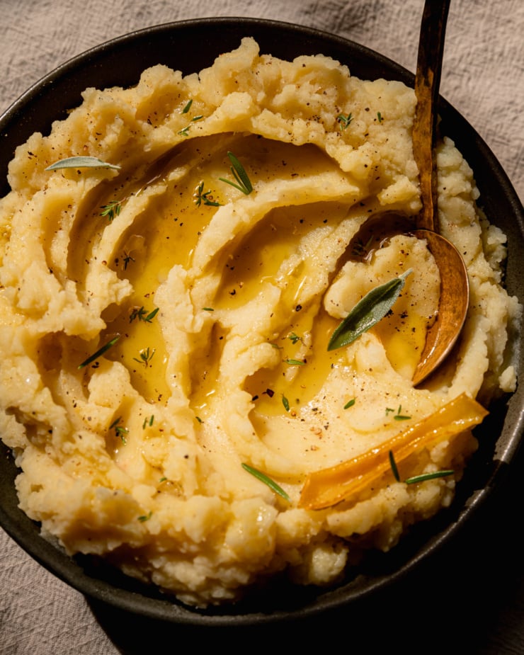 An up close, overhead shot of olive oil mashed potatoes in a bowl with swoops of olive oil on top and little leaves of herbs as well. A brass serving spoon is sticking out of the potatoes.