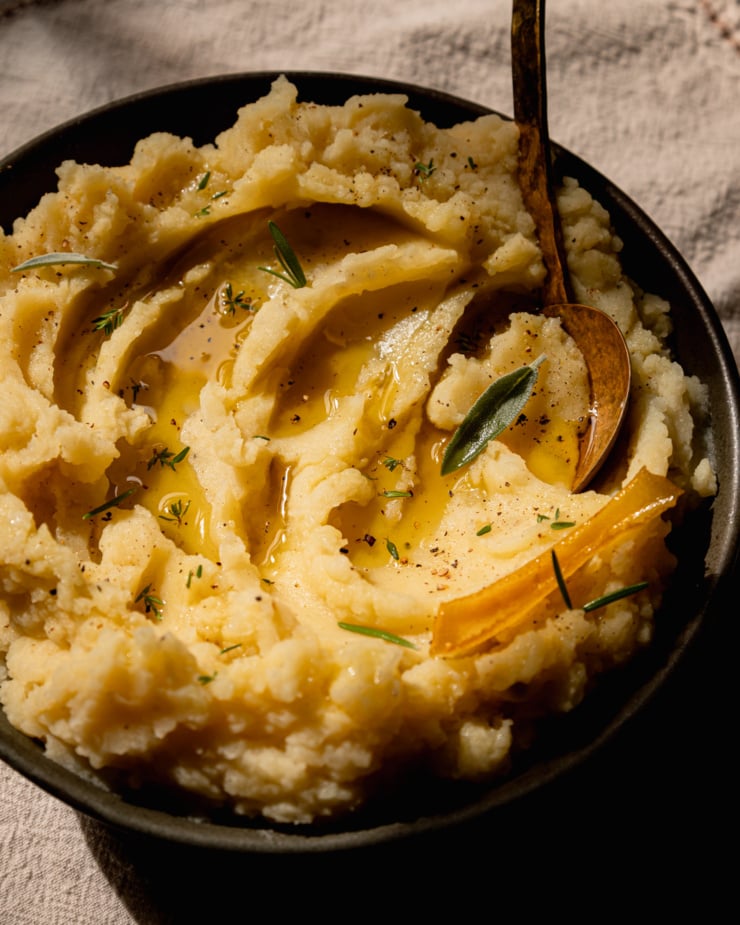 A 3/4 angle shot shows a bowl of creamy olive oil mashed potatoes with swoops of olive oil and whole herb leaves on top. A serving spoon is sticking out of the bowl.