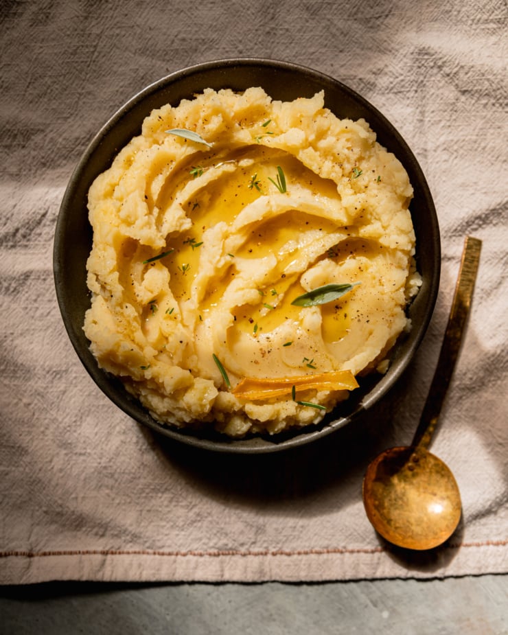An overhead shot of olive oil mashed potatoes in a bowl with swoops of olive oil on top and little leaves of herbs as well. A brass serving spoon is nearby.