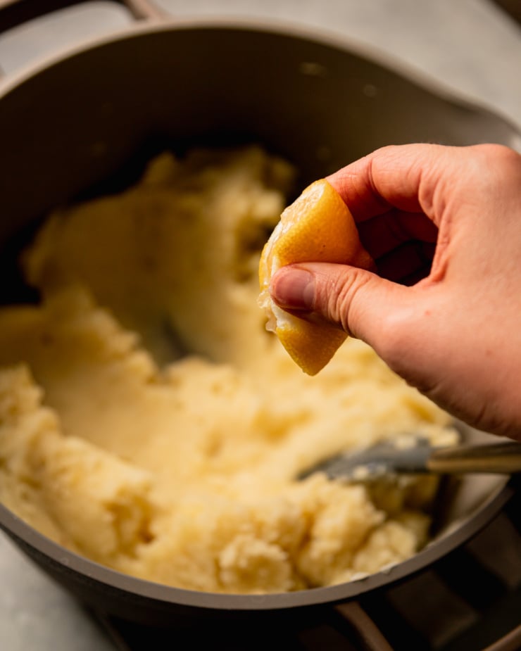 A slight 3/4 angle image shows. hand squeezing a lemon half into a pot of mashed potatoes.