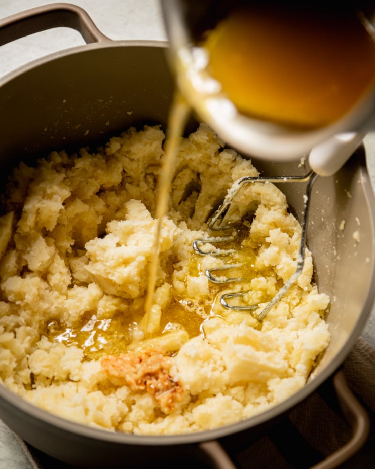 An overhead shot shows warm vegetable stock and olive oil being added to a pot of mashed potatoes.