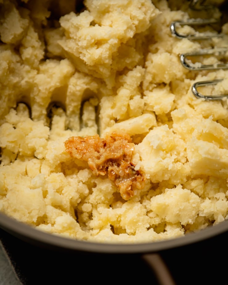 An overhead shot shows a clump of roasted garlic on top of a bunch of mashed potatoes in a pot.