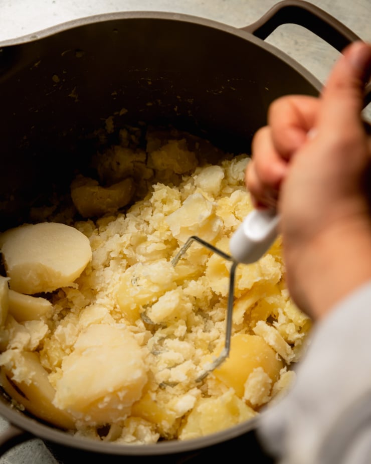 An overhead shot shows a hand using a masher to mash potatoes in a pot.