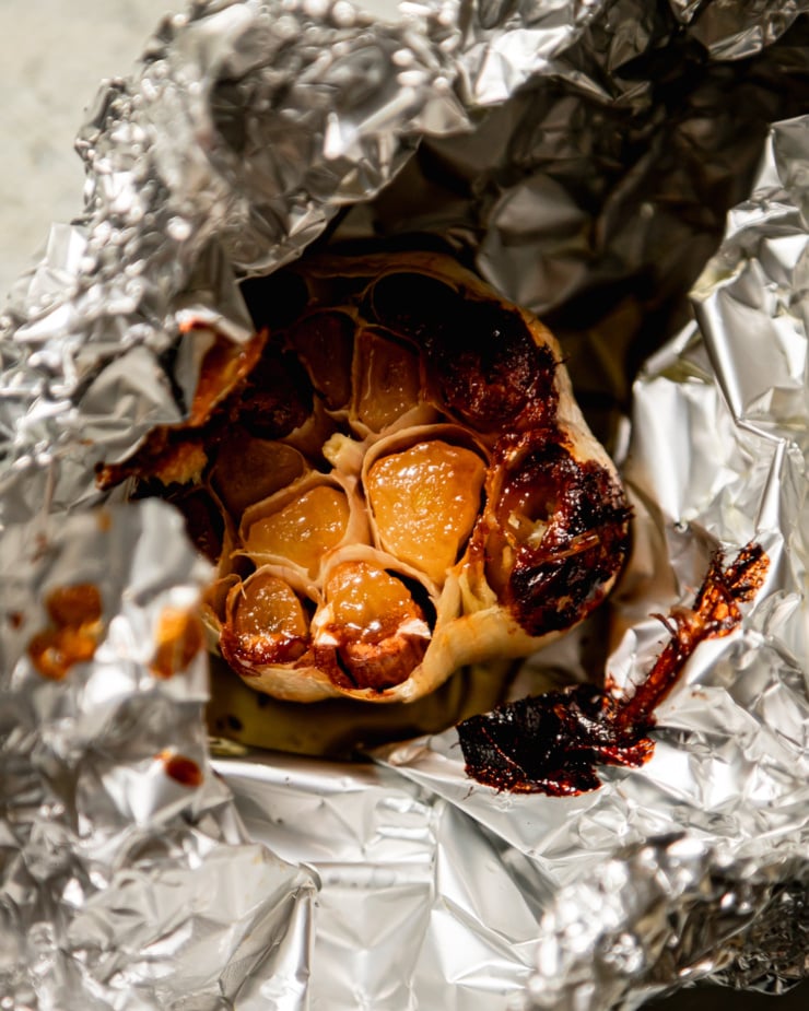 An up close, overhead shot shows a bulb of roasted garlic, still partially wrapped in aluminum foil.