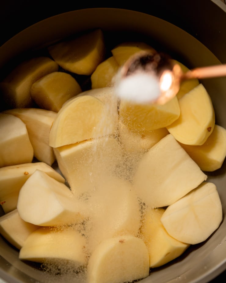 An overhead shot shows peeled and diced potatoes submerged in water in a pot. A spoon is sprinkling some salt into the pot.