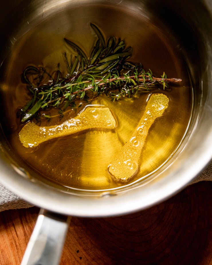 An overhead shot of lemon peel and herb sprigs sizzling in a pot of olive oil.