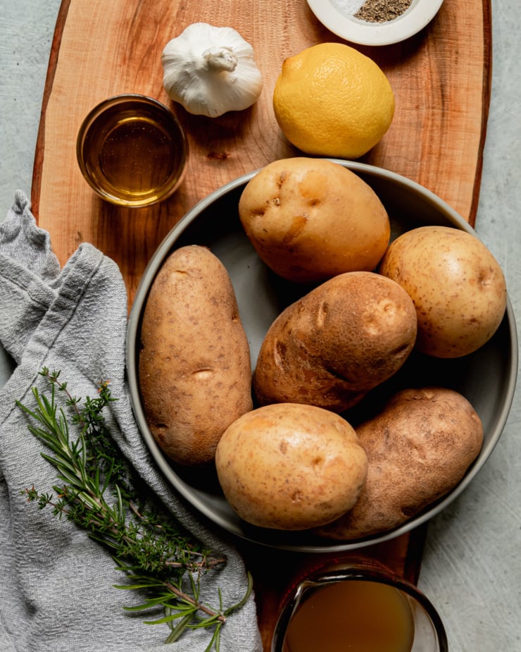 An overhead shot of ingredients used in olive oil mashed potatoes: yukon gold potatoes, russet potatoes, garlic, lemon, rosemary, thyme, olive oil, vegetable stock, salt, and pepper.