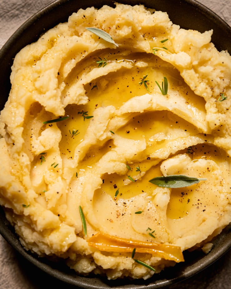 An up close, overhead shot of olive oil mashed potatoes in a bowl with swoops of olive oil on top and little leaves of herbs as well.