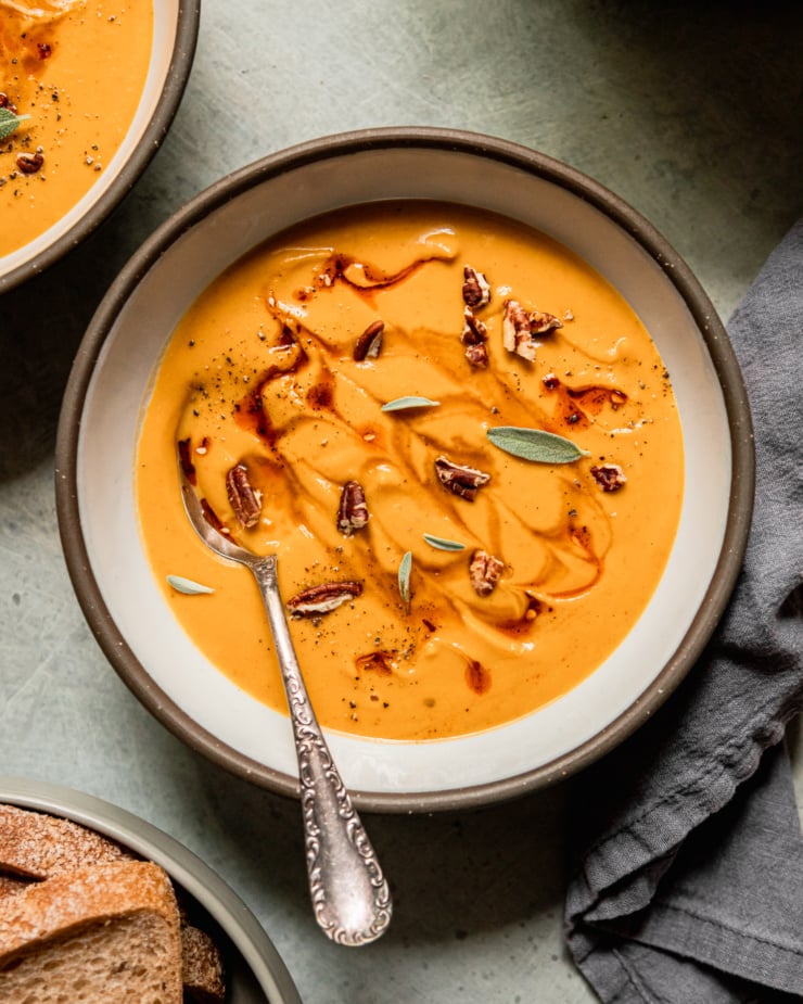 An overhead shot shows a bowl of vegan butternut squash soup garnished with small sage leaves, chopped pecans, and drizzles of maple syrup and chili crisp. A spoon is sticking out of the bowl