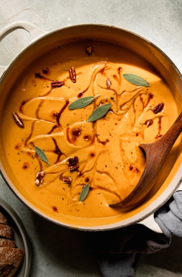 An overhead shot shows a pot of vegan butternut squash soup with apples and pecans. The puréed soup is garnished with maple syrup, chili crisp, small sage leaves, and chopped pecans. A wooden ladle is sticking out of the pot.