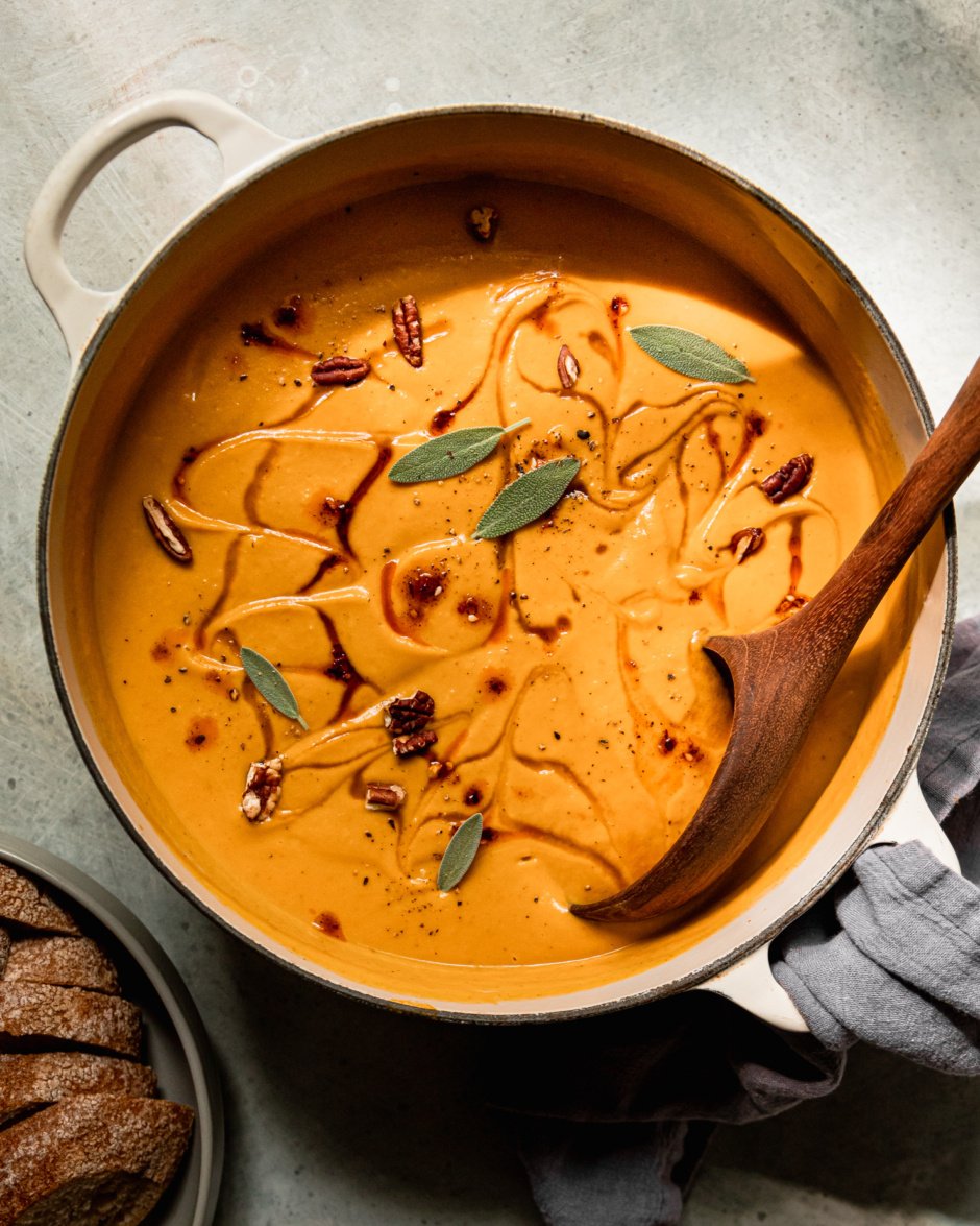 An overhead shot shows a pot of vegan butternut squash soup with apples and pecans. The puréed soup is garnished with maple syrup, chili crisp, small sage leaves, and chopped pecans. A wooden ladle is sticking out of the pot.