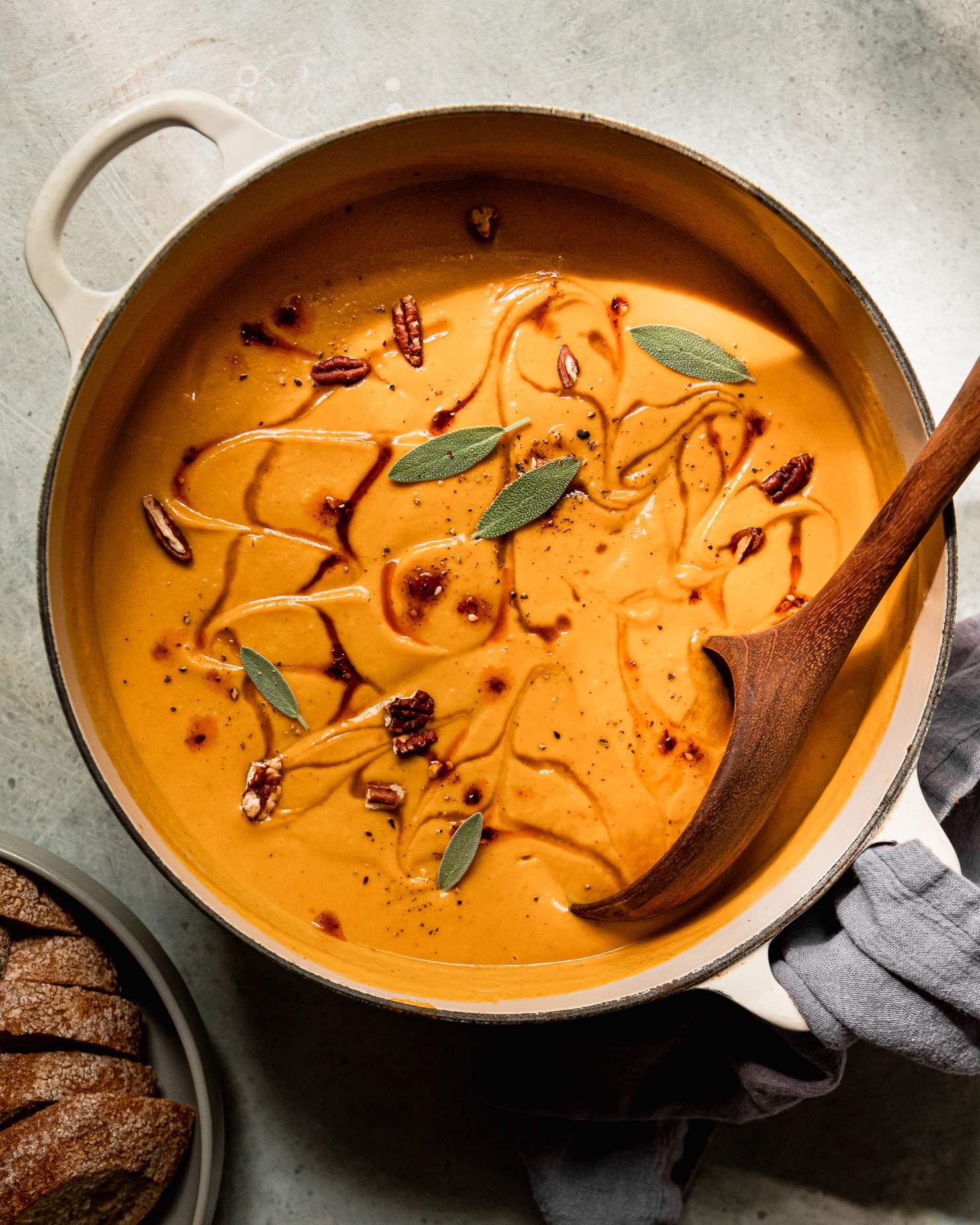 An overhead shot shows a pot of vegan butternut squash soup with apples and pecans. The puréed soup is garnished with maple syrup, chili crisp, small sage leaves, and chopped pecans. A wooden ladle is sticking out of the pot.