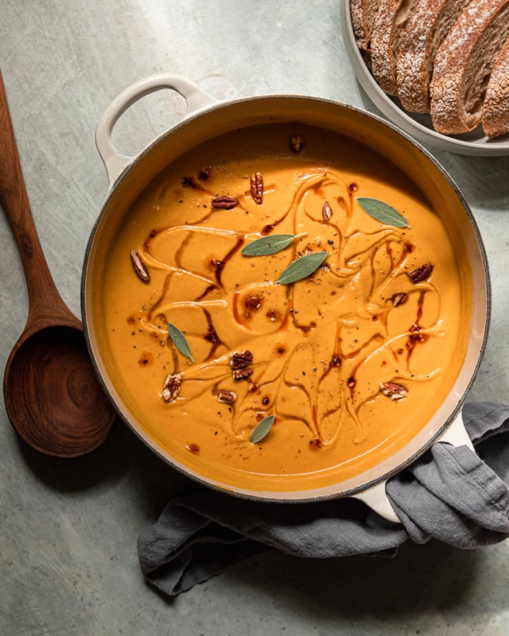 An overhead shot shows a pot of vegan butternut squash soup with apples and pecans. The puréed soup is garnished with maple syrup, chili crisp, small sage leaves, and chopped pecans.