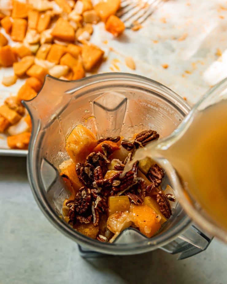 An overhead shot shows vegetable stock being poured into a blender pitcher that is filled with roasted butternut squash, onions, apples, and toasted pecans