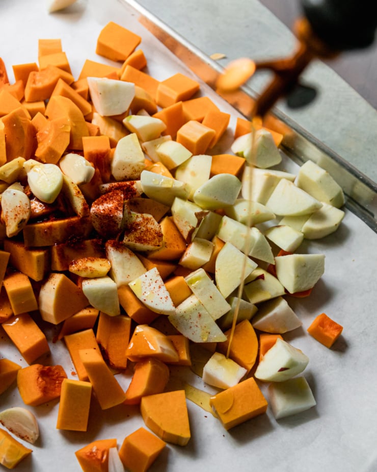 An overhead shot shows olive oil being poured on top of cubes butternut squash, chopped apples, chopped onions, garlic, and ginger, Ground chilies are sprinkled around as well. All is contained on a baking sheet.
