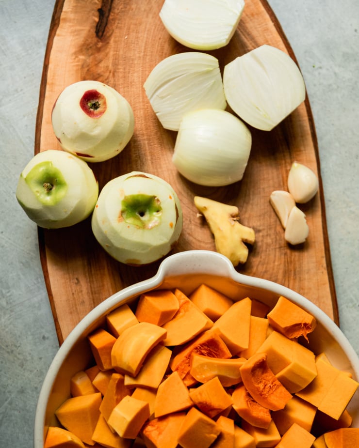 An overhead shot shows chopped butternut squash, peeled apples, peeled and halved onions, peeled garlic cloves, and a peeled chunk of ginger.