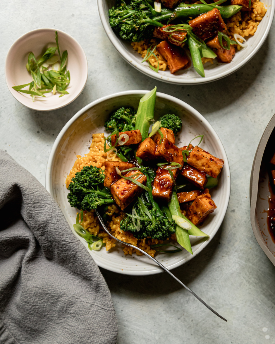 An overhead shot shows two spicy maple tofu bowls with golden garlic rice as a base and steamed broccolini. The bowls are topped with sliced green onions.