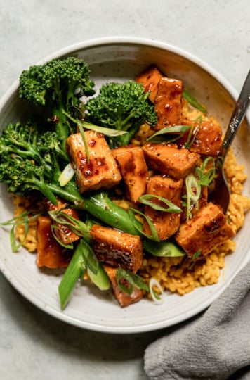 An overhead shot shows a spicy maple tofu bowl with golden garlic rice as a base and a few stalks of steamed broccolini. The bowl is topped with sliced green onions.