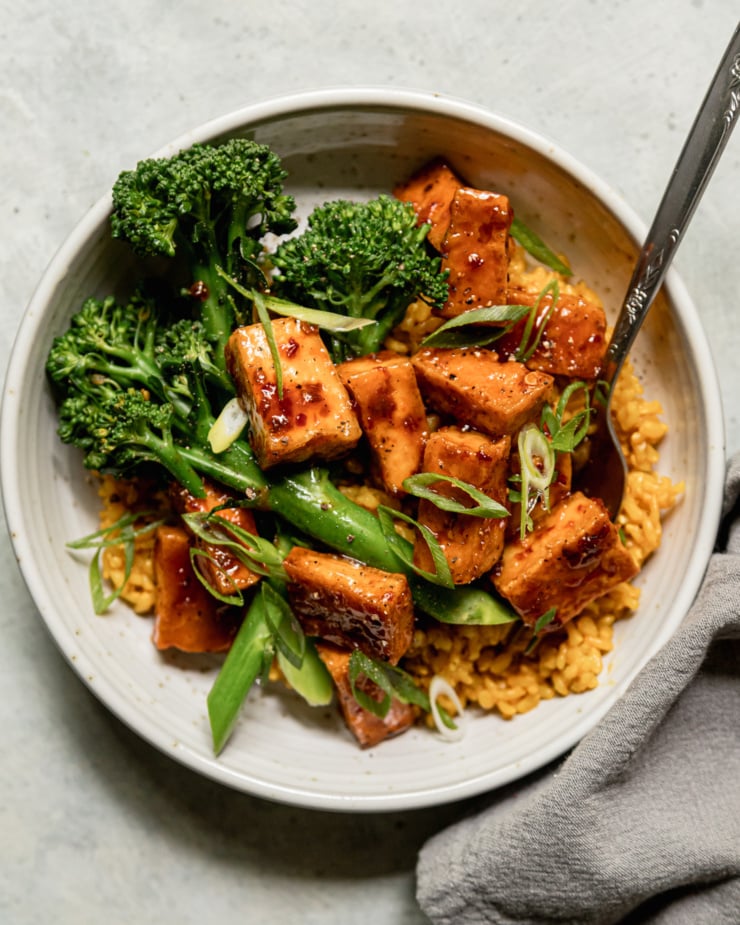 An overhead shot shows a spicy maple tofu bowl with golden garlic rice as a base and a few stalks of steamed broccolini. The bowl is topped with sliced green onions.