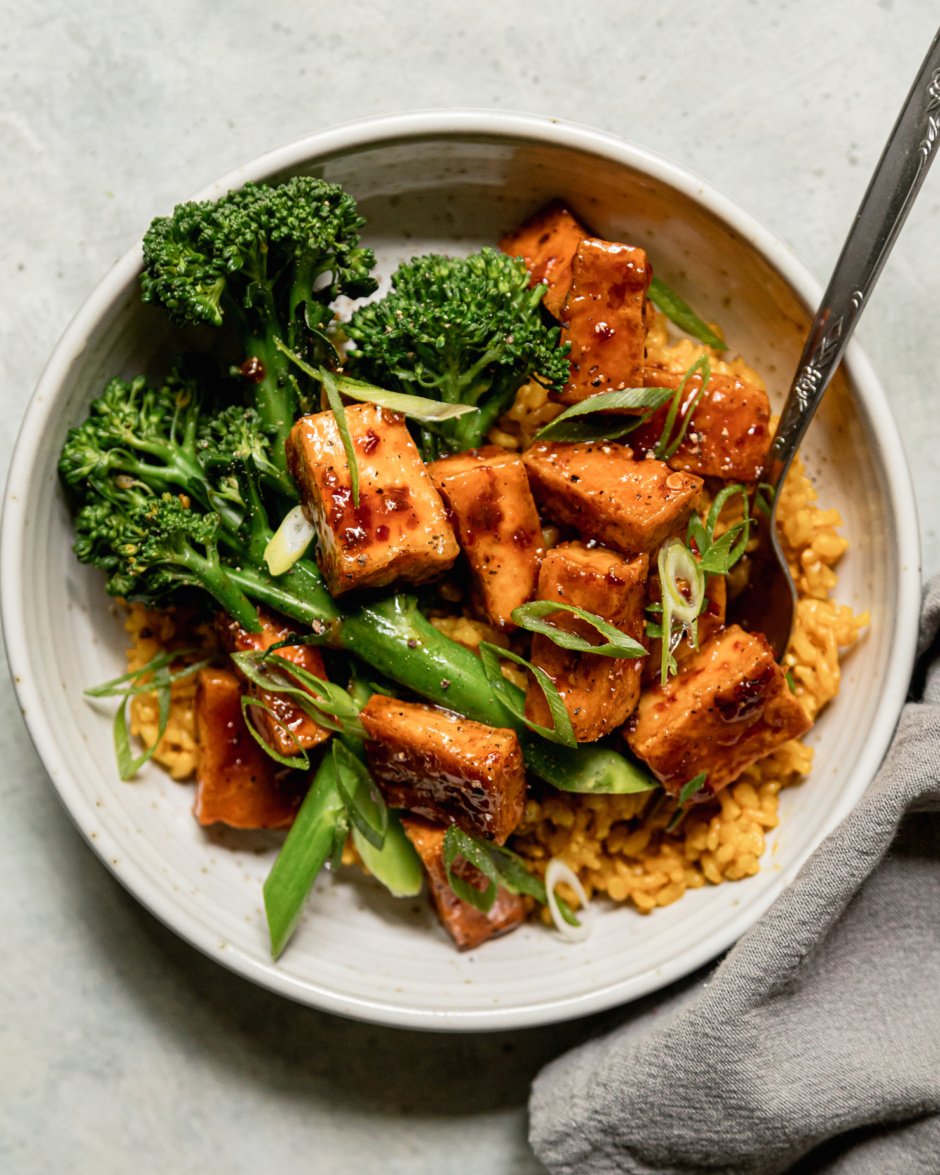 An overhead shot shows a spicy maple tofu bowl with golden garlic rice as a base and a few stalks of steamed broccolini. The bowl is topped with sliced green onions.