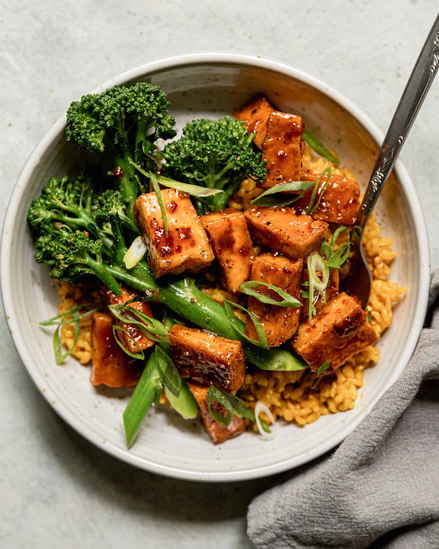 An overhead shot shows a spicy maple tofu bowl with golden garlic rice as a base and a few stalks of steamed broccolini. The bowl is topped with sliced green onions.