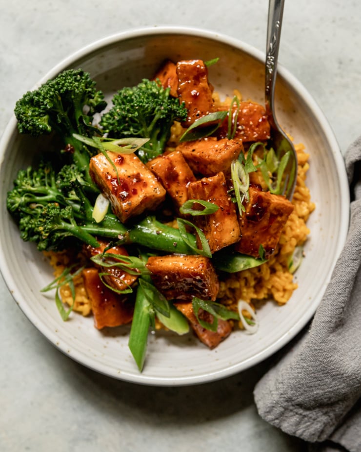 An overhead shot shows a spicy maple tofu bowl with golden garlic rice as a base and a few stalks of steamed broccolini. The bowl is topped with sliced green onions.
