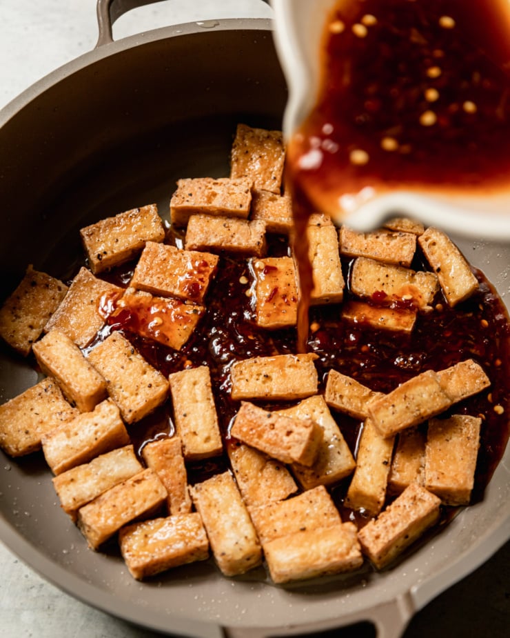 A 3/4 angle shot shows fried tofu in a skillet with a spicy maple sauce being poured in.